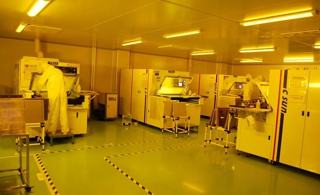 Workers in cleanroom suits operate photolithography machines under yellow light in a PCB manufacturing facility, ensuring precise circuit board production.